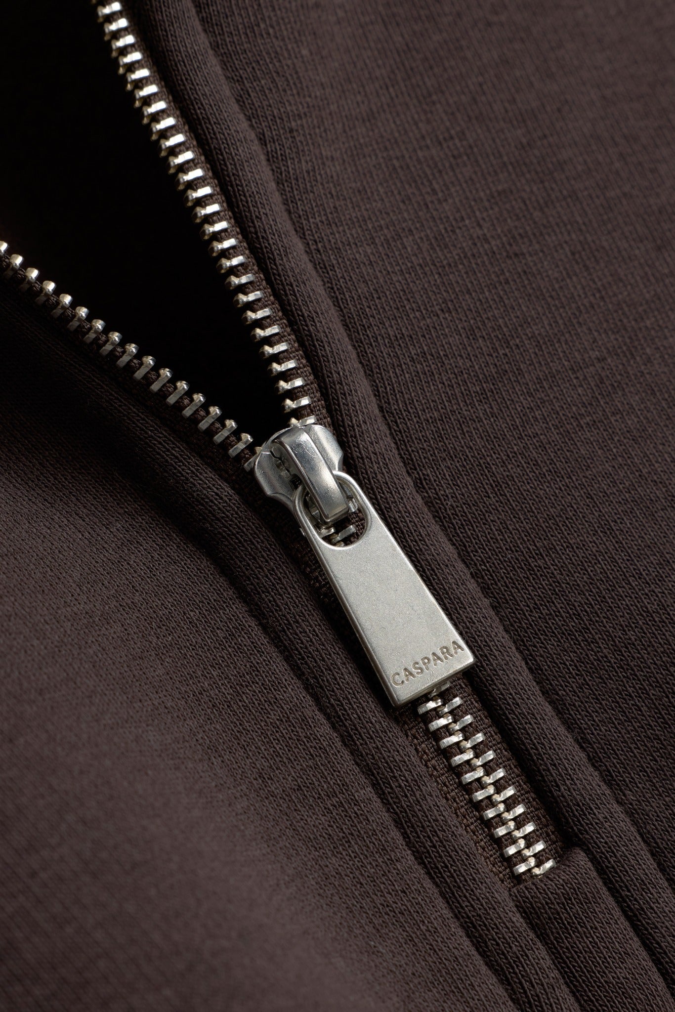 Close-up of the silver zipper pull engraved with CASPARA on an espresso brown quarter-zip sweatshirt.