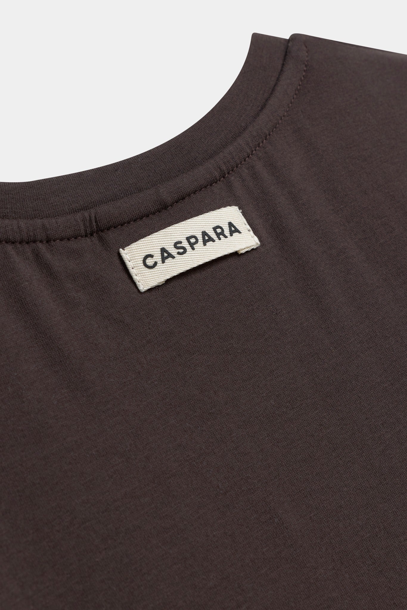 Close-up of the woven CASPARA logo label on the neckline of an espresso baby tee.