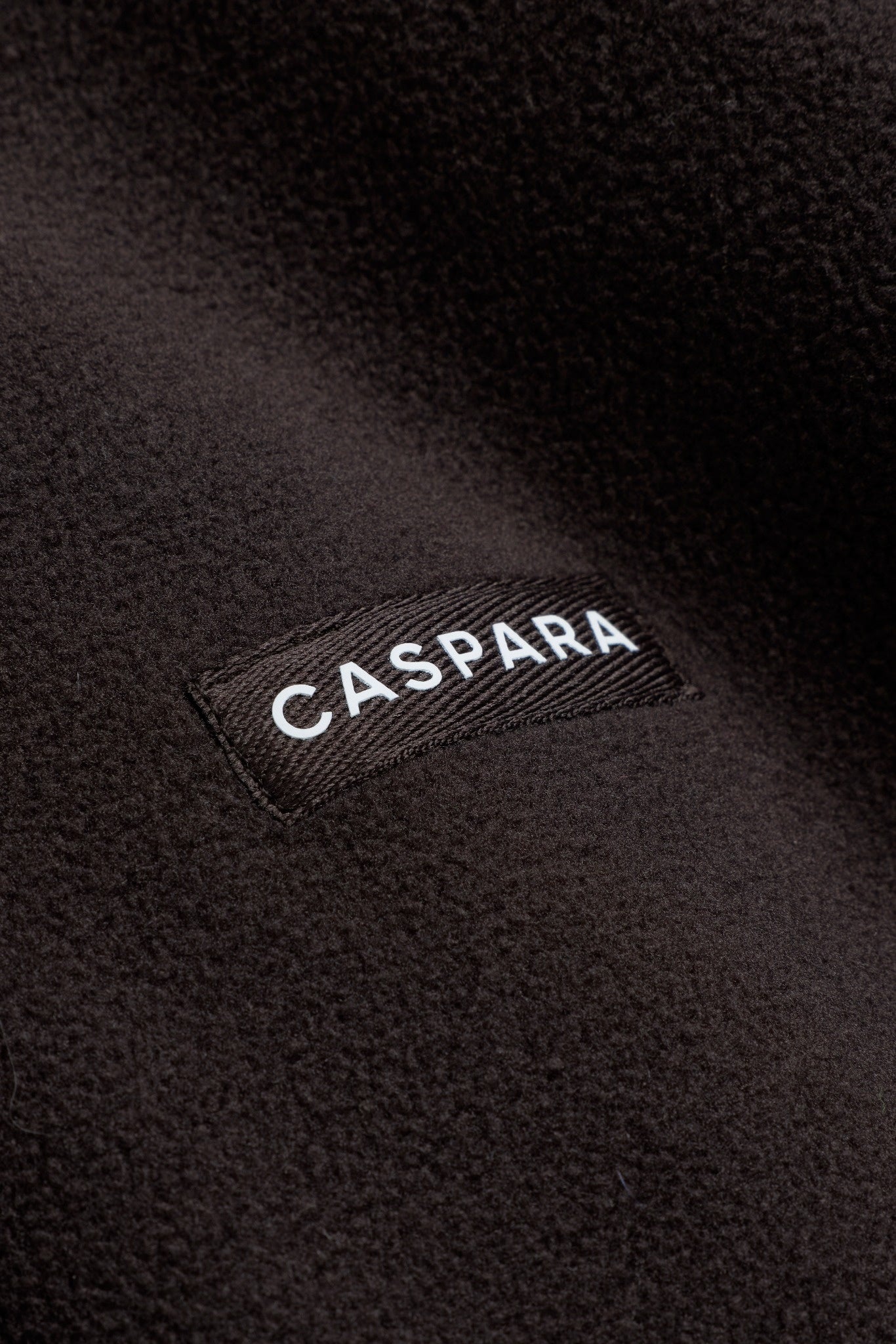 Close-up of the CASPARA logo label on an espresso brown polar fleece pullover.
