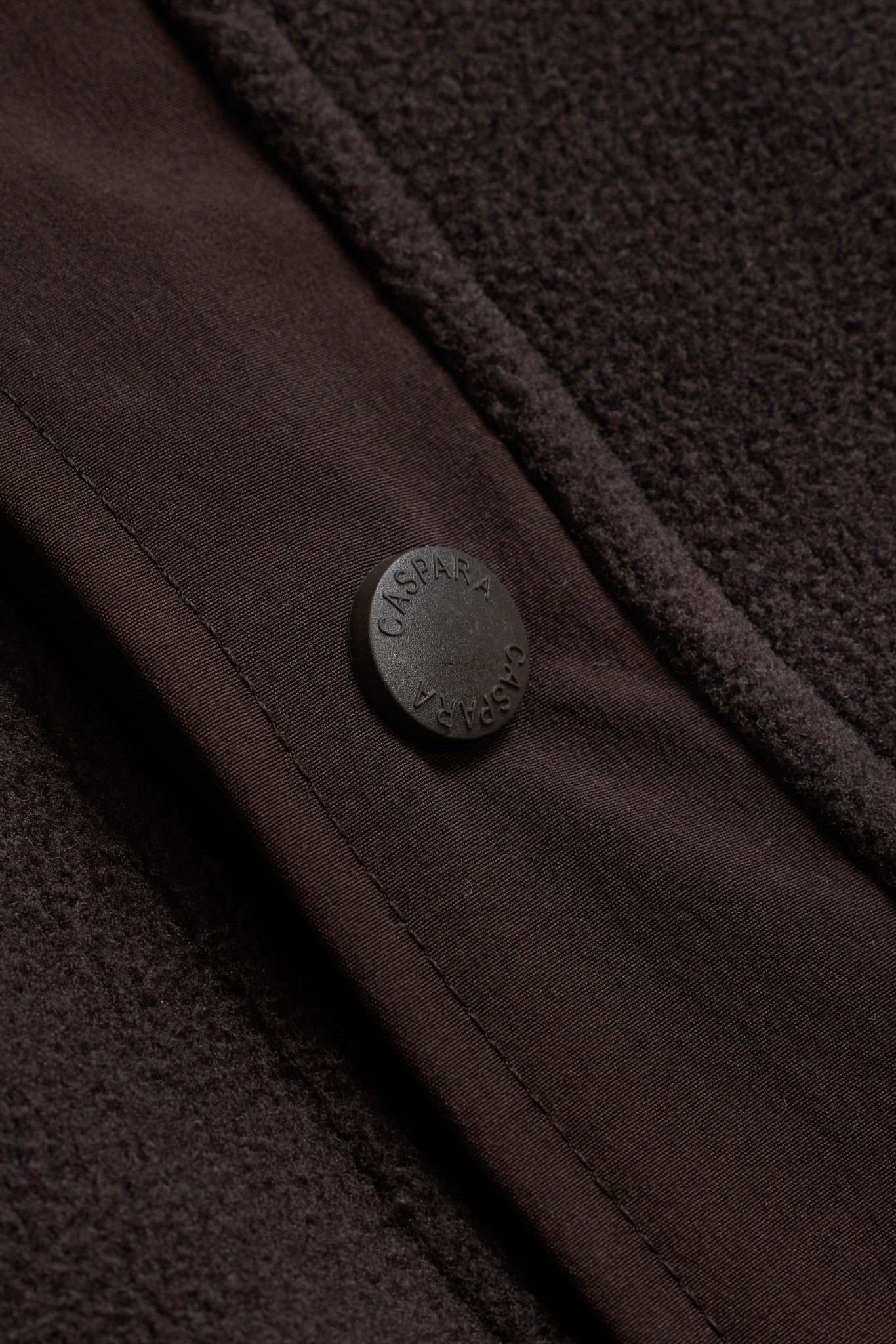 Close-up of an espresso brown fleece pullover featuring an embossed CASPARA snap button.