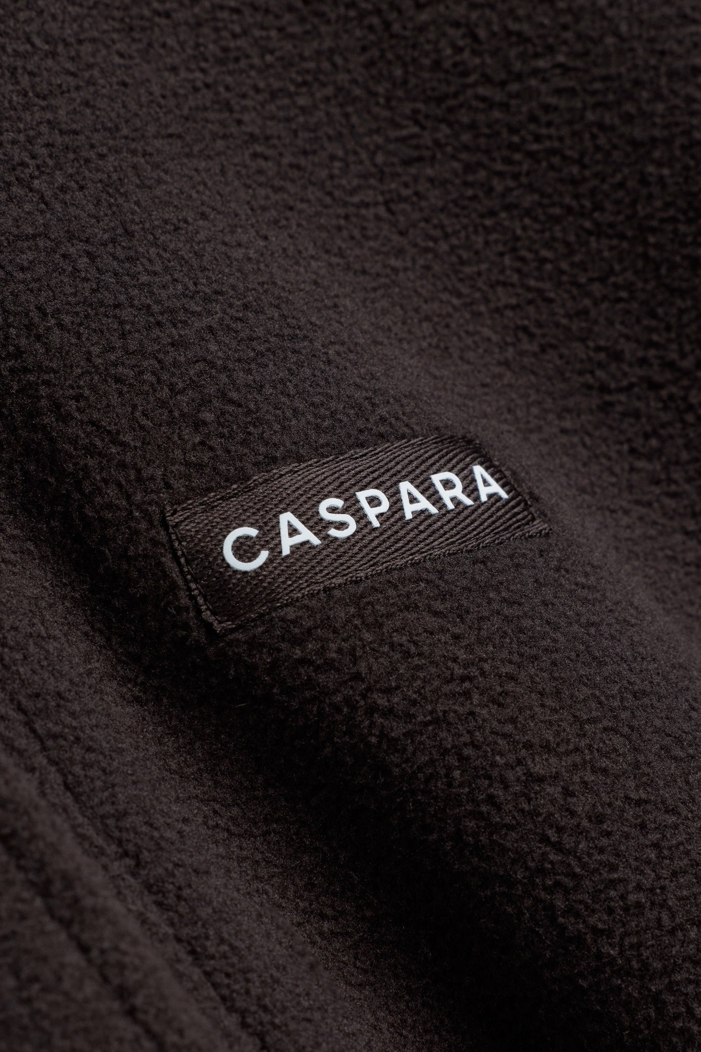 Close-up of the CASPARA logo label on a cocoa bean polar fleece pullover.