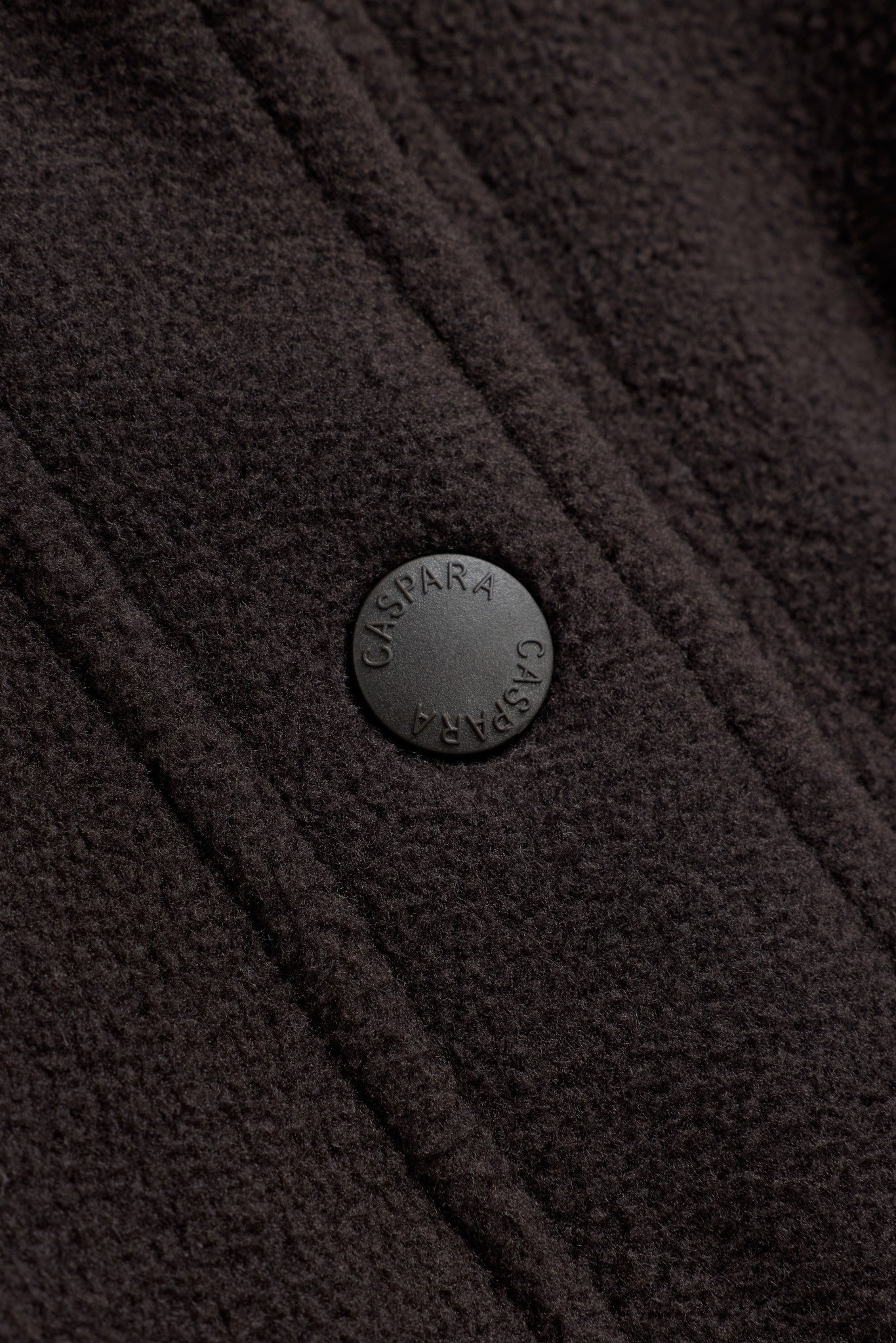 Close-up of an engraved CASPARA snap button on a cocoa bean polar fleece pullover.