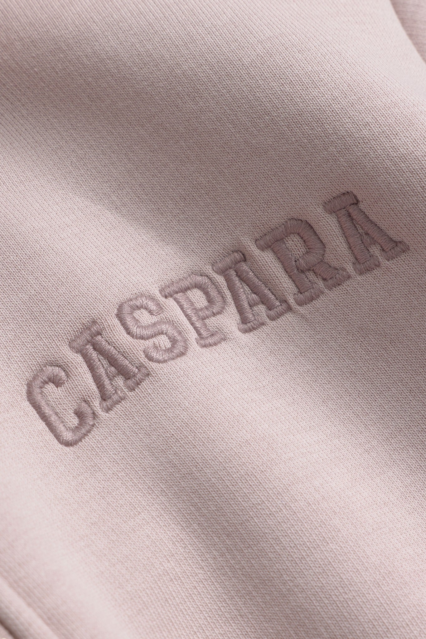 Close-up of the tonal embroidered CASPARA logo on a dusty pink zip hoodie.