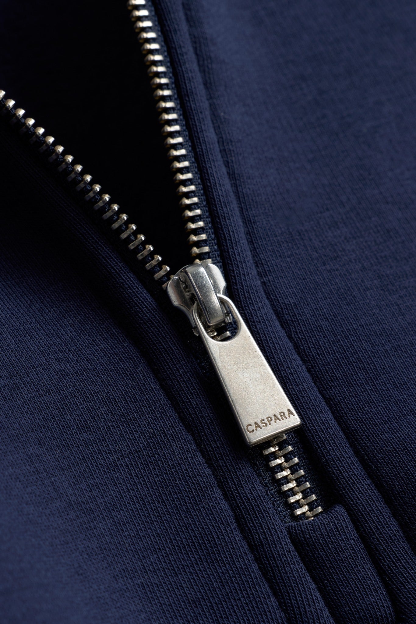 Close-up of the silver zipper pull engraved with CASPARA on a navy sweatshirt.