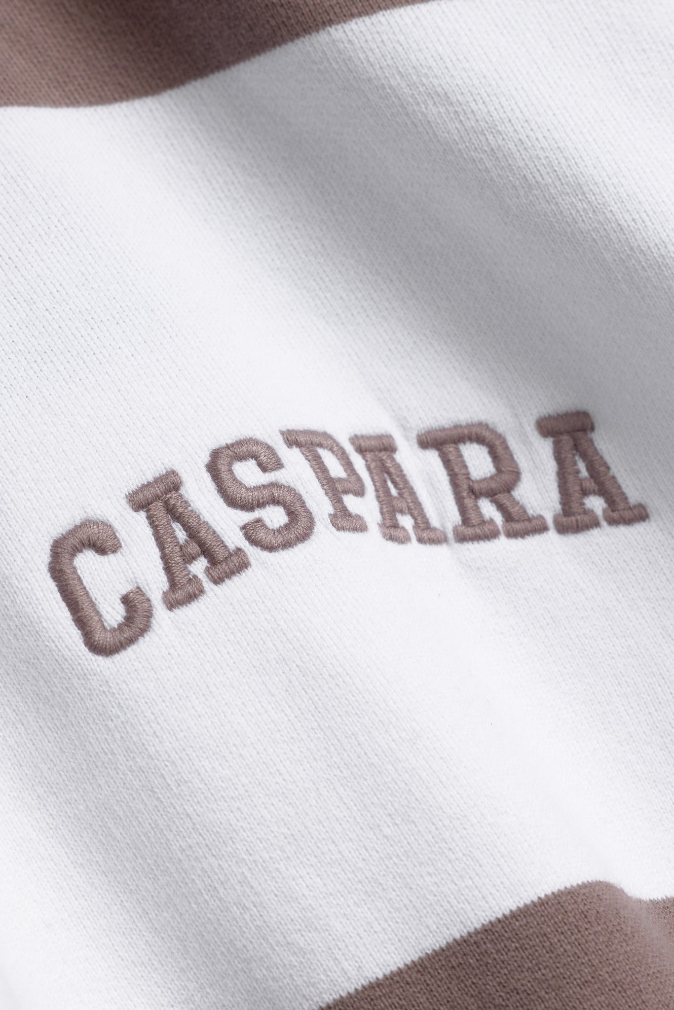 Close-up of embroidered CASPARA logo on a walnut and white striped cotton rugby shirt.