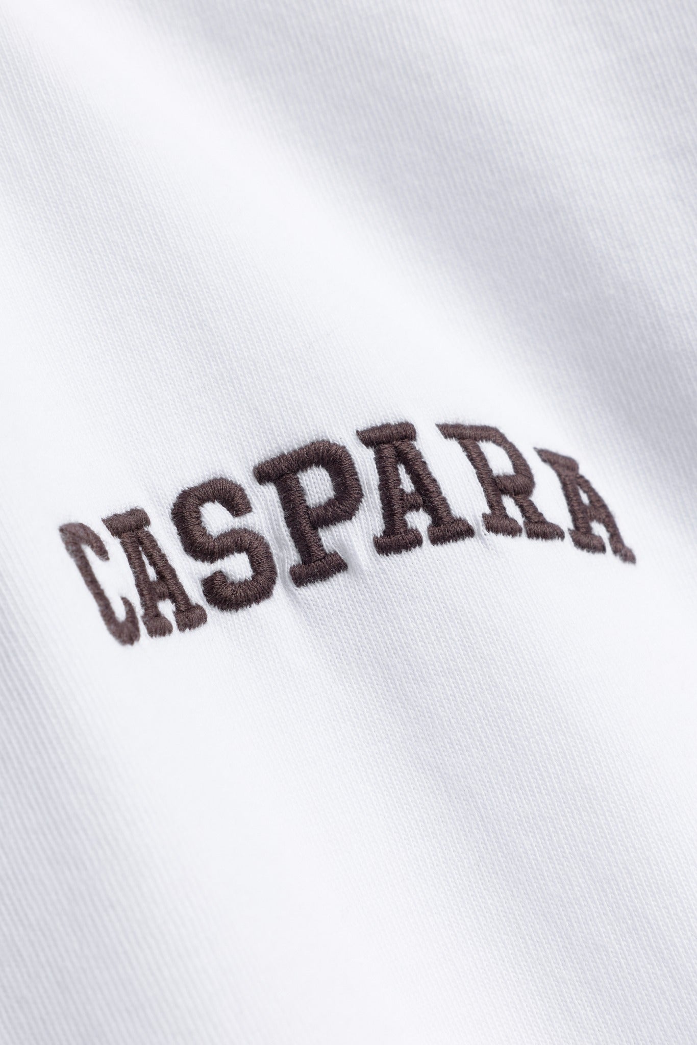 Close-up of espresso embroidered CASPARA logo on a white cotton t-shirt.