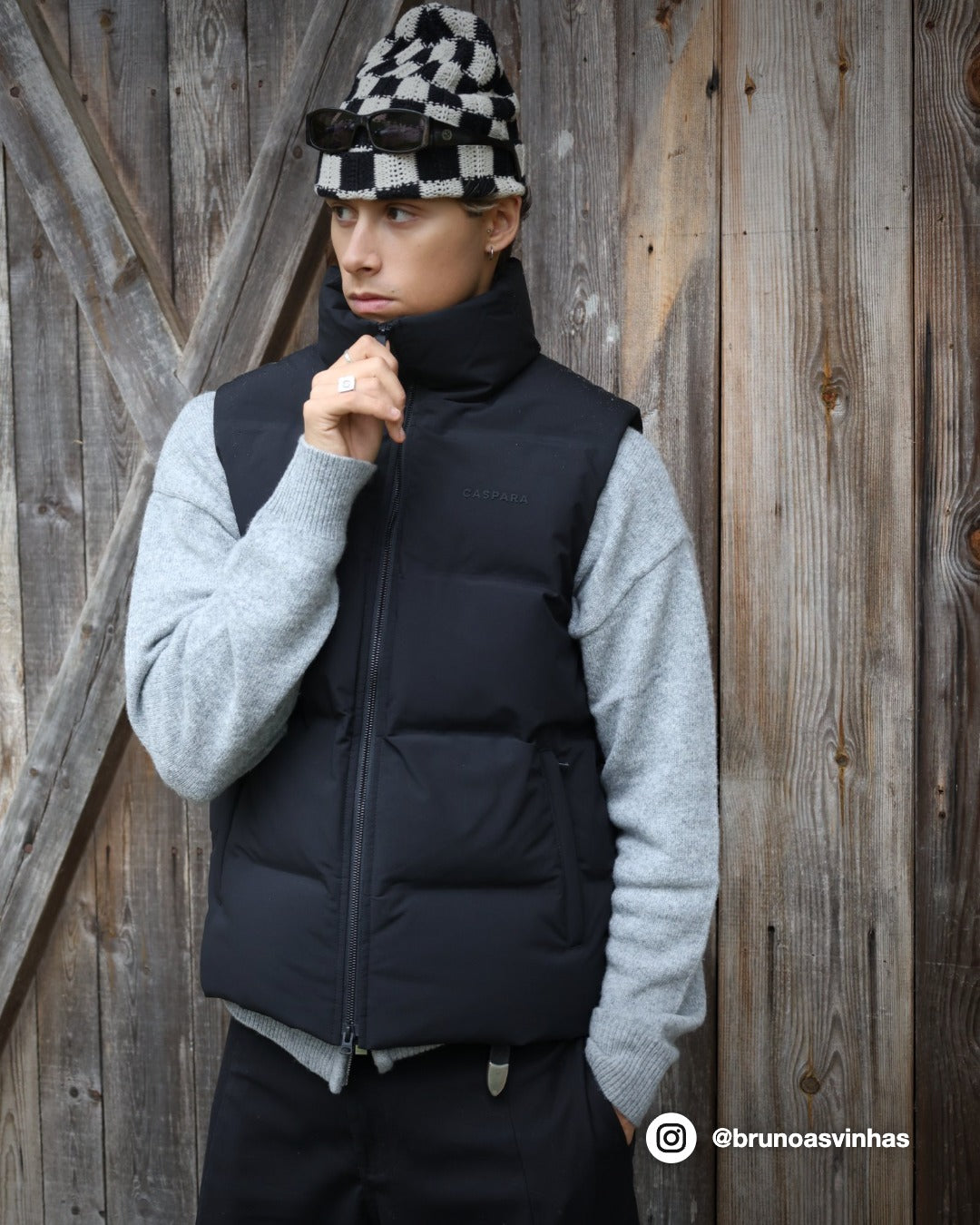 Man wearing a black CASPARA Boreal puffer vest over a grey sweater against a wooden wall.