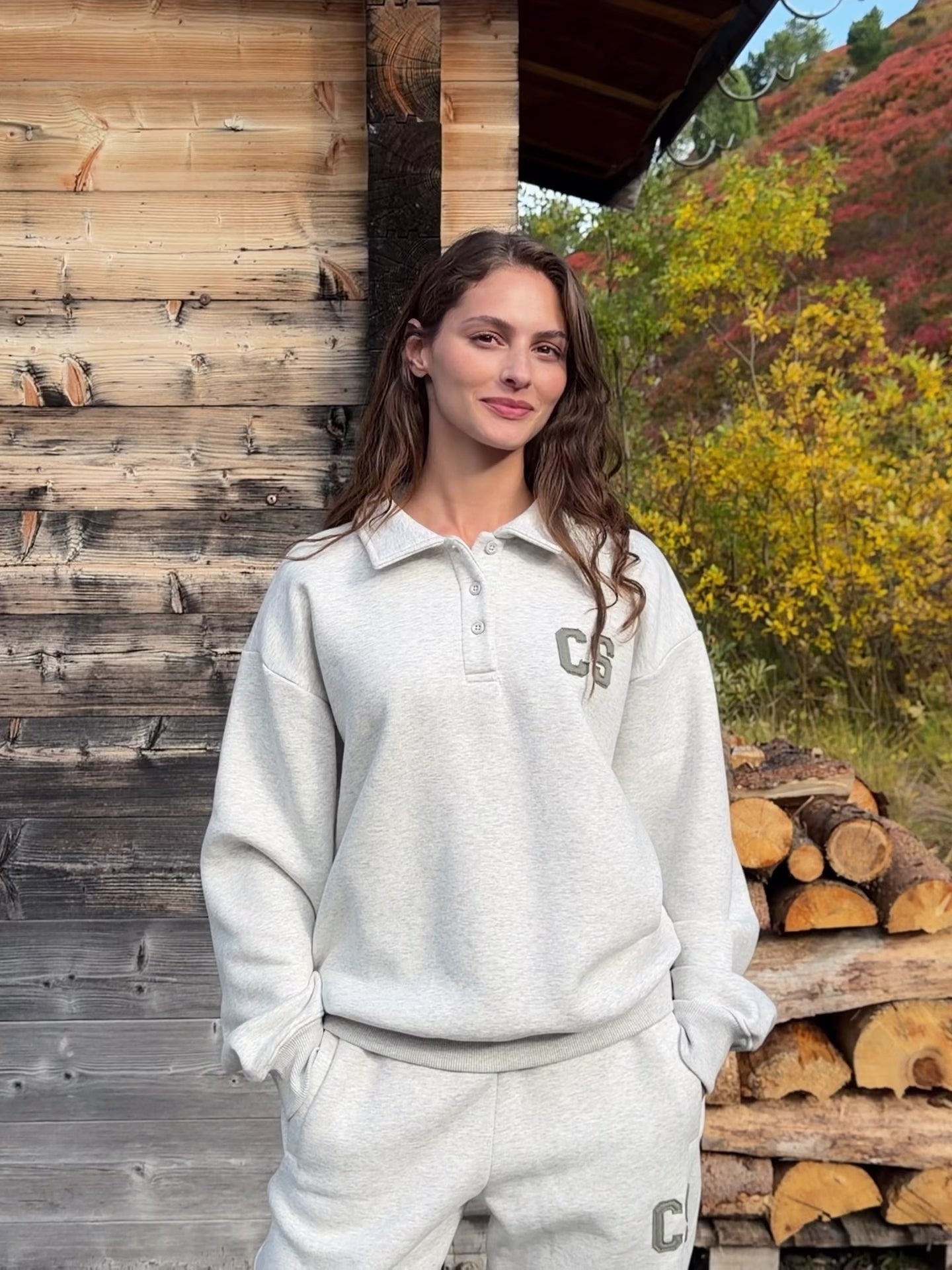 Woman wearing a grey melange CASPARA CS rugby shirt next to a wooden cabin.