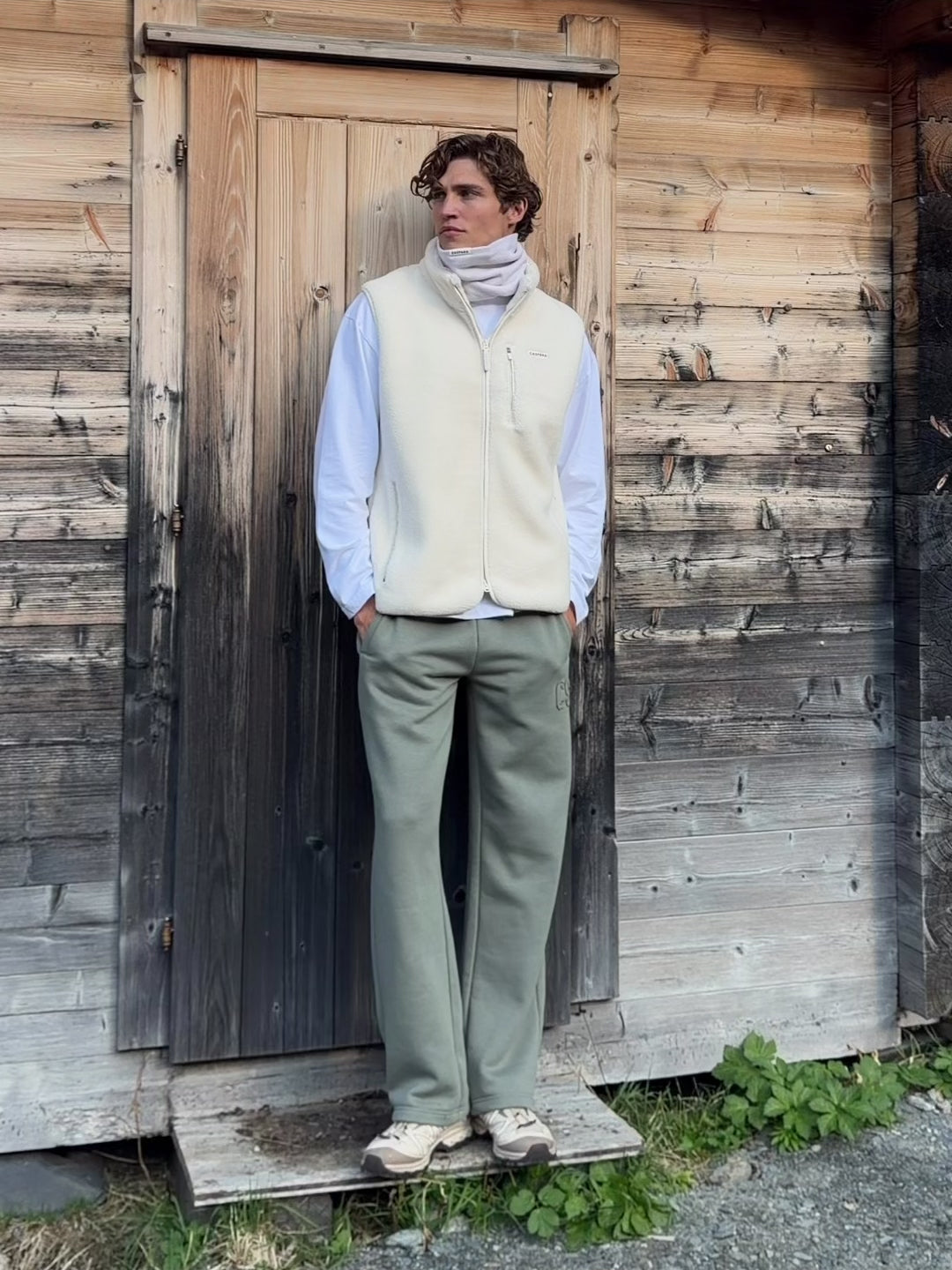 Man wearing a creme CASPARA Cloudfleece vest outside a wooden cabin.
