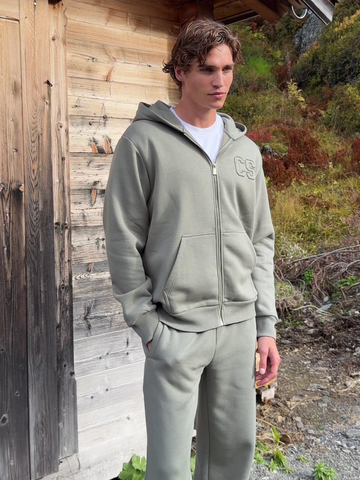 Man wearing a khaki CASPARA CS zip hoodie standing outside a wooden cabin.