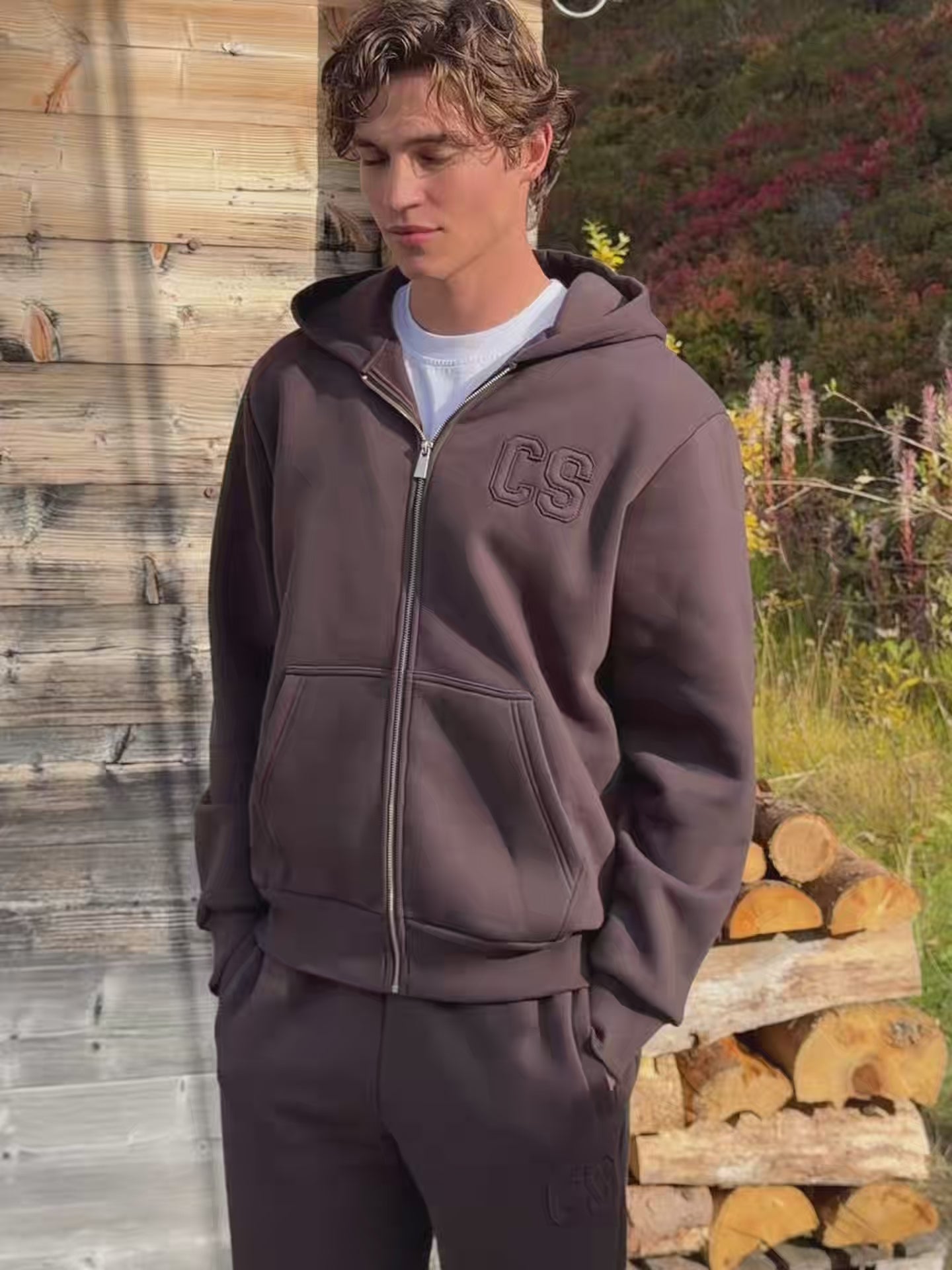 Man wearing an espresso CASPARA CS zip hoodie outdoors near a wooden cabin.
