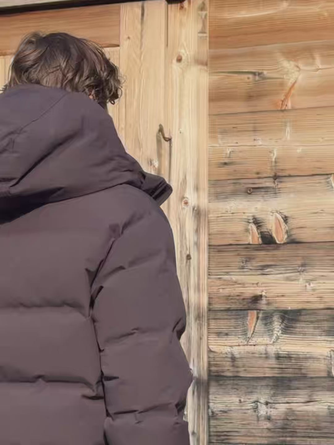 Man wearing a cocoa bean CASPARA Boreal puffer jacket outside a wooden cabin.