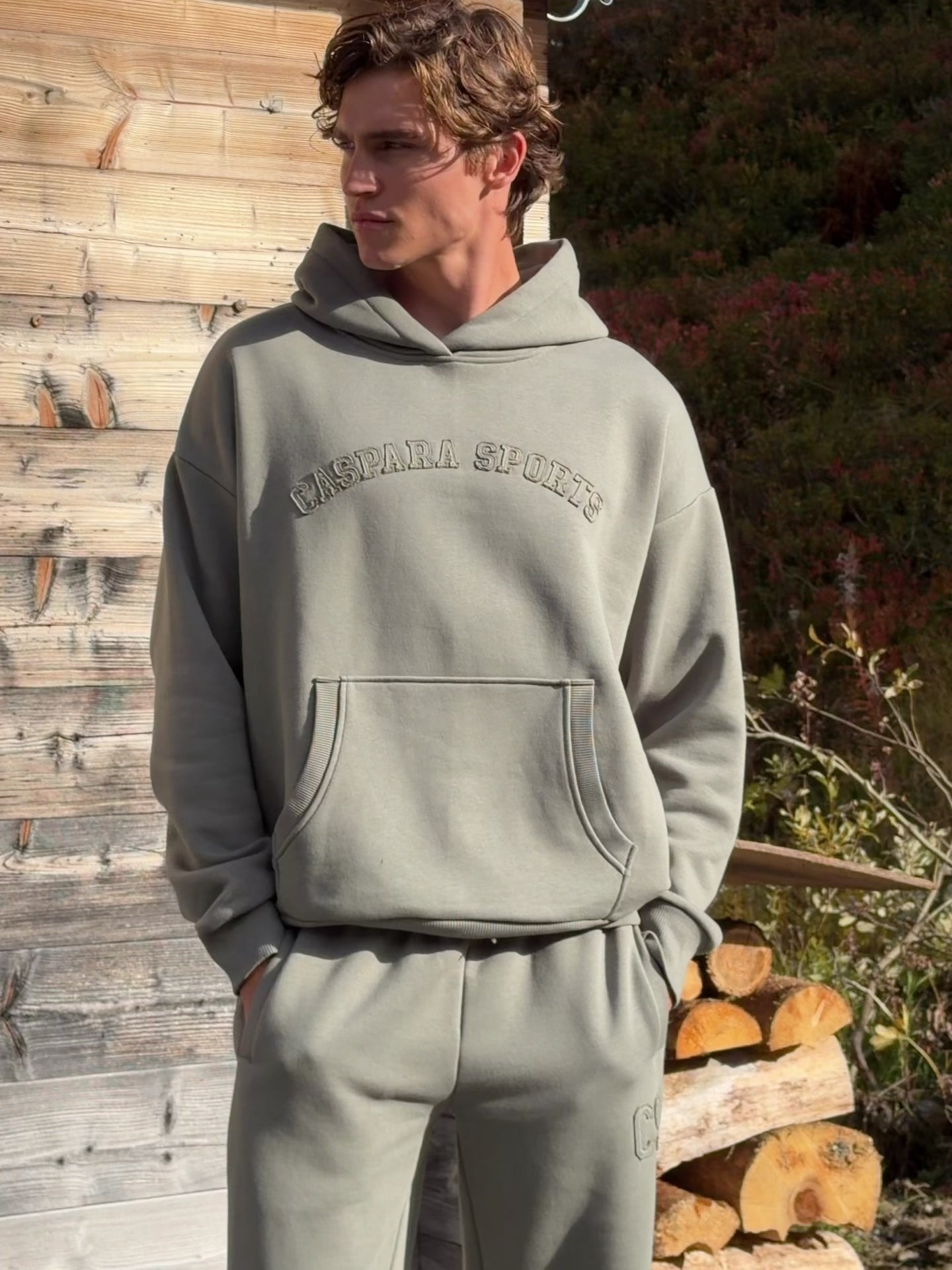 Man wearing a khaki CASPARA CS hoodie standing outside a wooden cabin.